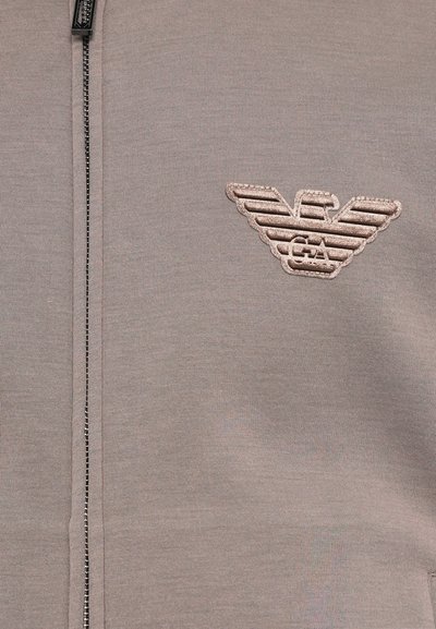 Pilkas zip-up džemperis su minkšta tekstūra ir išsiuvinėtų logotipu, kuris turi sparnų dizainą kairiajame krūtinės srityje.