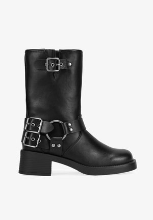Bottine en cuir noir à bout rond, avec deux boucles sur les côtés et un détail de sangle circulaire. Semelle texturée et talon empilé.