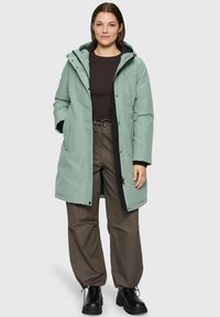 Femme portant un parka à capuche vert clair, un haut à manches longues marron foncé, un pantalon marron ample et des bottines noires, se tenant avec les mains dans les poches.