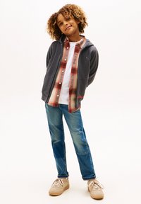 Grå zip-up hættetrøje over en ternet skjorte med rød- og cremetoner, hvid T-shirt, blå denim jeans og beige sneakers.