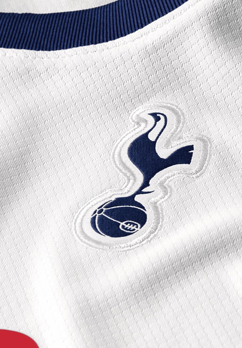 Biały strój sportowy z teksturowanej tkaniny, granatowym prążkowanym kołnierzykiem oraz wyraźnym, granatowym haftowanym logo przedstawiającym ptaka nad piłką.