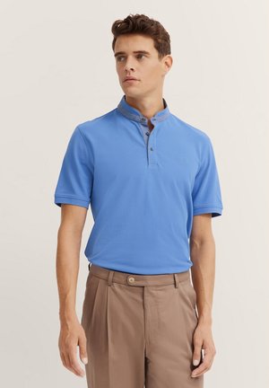 Blaues Poloshirt mit Stehkragen, das einen kontrastierenden Stoff am Halsausschnitt und kurze Ärmel aufweist. Hellbraune Hose vervollständigt das Outfit.