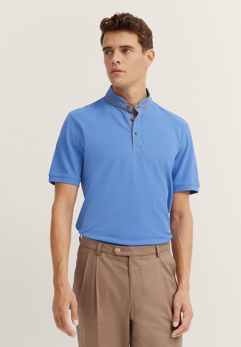 Blaues Poloshirt mit Stehkragen, das einen kontrastierenden Stoff am Halsausschnitt und kurze Ärmel aufweist. Hellbraune Hose vervollständigt das Outfit.