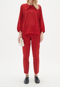 Czerwona bluzka z bufkami i skręconym detalem, zestawiona z dopasowanymi czerwonymi spodniami. Czarne buty na sznurówki, trzymająca kremową kopertówkę.