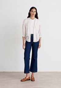 White, lightweight, button-up blouse with gathered detailing, paired with dark blue wide-leg jeans and brown moccasin-style shoes.