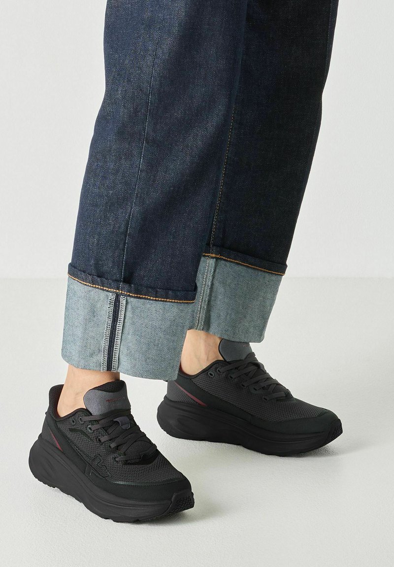 Jambes portant un jean denim foncé retroussé et des baskets noires à lacets, debout sur un sol clair uni.