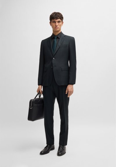 Costume vert foncé avec une finition texturée, coupe ajustée, blazer à un bouton, pantalon assorti et un sac en cuir noir. Des chaussures de ville noires terminent le look.