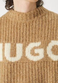Pull en tricot confortable couleur beige avec le logo "HUGO" en crème bien en évidence. Présente une texture côtelée et un design à col montant.