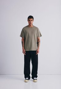 Maglietta oversize verde oliva con maniche corte e collo rotondo, abbinata a pantaloni neri a gamba larga e sneakers nere e bianche. Tessuto strutturato.