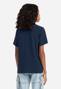 Young person with curly hair wears a plain navy blue short-sleeve shirt and light blue cargo jeans, facing away against a white background.