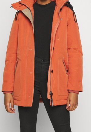 Person trägt eine orangefarbene Kapuzenparka mit Fronttaschen über einem schwarzen Hemd und schwarzen Hosen und steht vor einem neutralen Hintergrund.