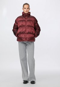 Blizganti bordo pūkinė striukė su aukštu apykakle, užtrauktuku priekyje ir įribuotomis rankovėmis, derinama su šviesiai pilkais plačiais džinsais ir juodais batais.