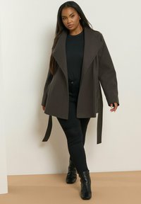 Donkergrijze wikkeljas met een grote kraag en strikceintuur, gecombineerd met een zwart top en broek, gedragen met zwarte enkellaarsjes, tegen een neutrale achtergrond.