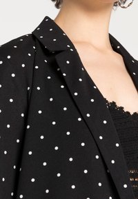 Schwarzer Blazer mit weißen Polka-Dots, ausgestattet mit einem Reverskragen und glatter Textur. Der Spitzenrand eines Unterkleidungsstücks ist teilweise sichtbar.