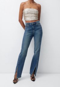 Top court sans bretelles en sequins dorés, associé à un jean en denim bleu clair taille haute avec des fentes sur les côtés et des sandales à talons noires.