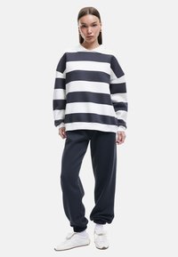 Pullover in Marineblau und weißen horizontalen Streifen, mit einem kragenlosen Ausschnitt und langen Ärmeln. Kombiniert mit dunklen Jogginghosen und weißen Sneakern.