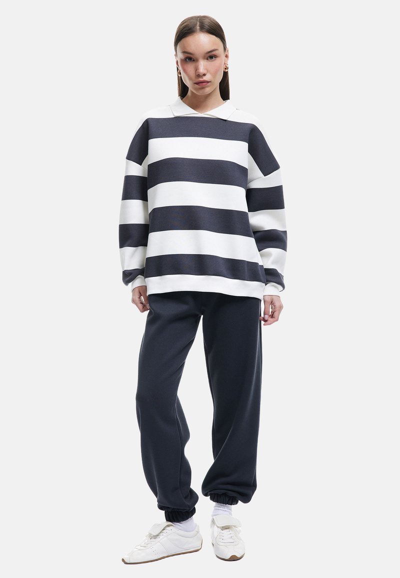 Pullover in Marineblau und weißen horizontalen Streifen, mit einem kragenlosen Ausschnitt und langen Ärmeln. Kombiniert mit dunklen Jogginghosen und weißen Sneakern.