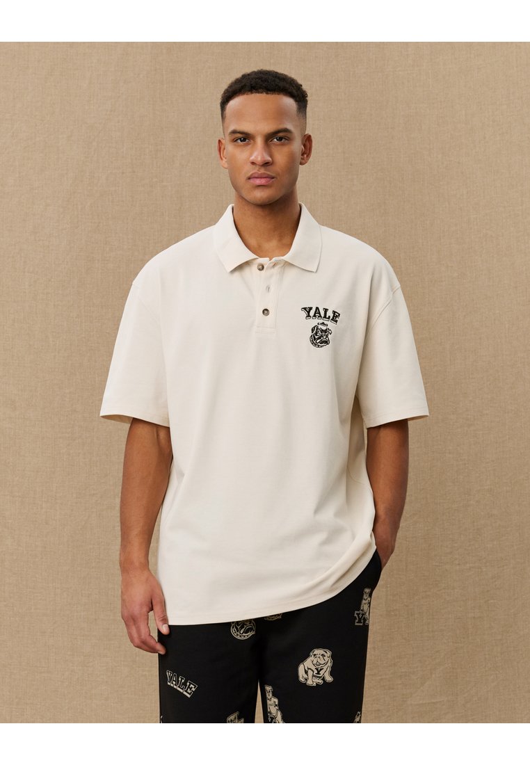 Polo shirt crem din bumbac, cu guler, având un logo Yale și un grafic pe pieptul stâng. Modelul pozează pe un fundal neutru.