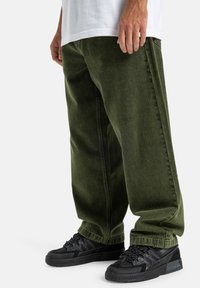 Zielone dżinsy o luźnym kroju, z bocznymi kieszeniami i lekko wyblakłą fakturą. W zestawie z czarnymi, teksturowanymi sneakersami.