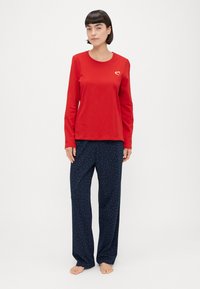 Rotes langärmliges Oberteil mit einem kleinen goldenen Herzlogo, kombiniert mit navyblauen Schlafanzughosen, die ein verstreutes, mehrfarbiges Punktmuster aufweisen.