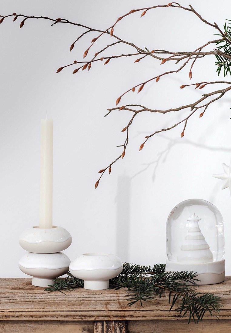 Portacandela in ceramica bianca con una candela alta color crema, un portacandela tondo impilato e una cupola di vetro con un albero, su una superficie di legno.
