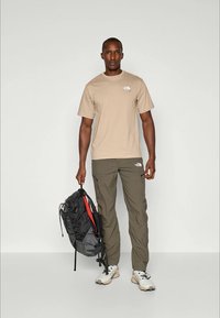 Beige bomulls t-shirt med logotyp, olivgröna cargobyxor, svart och röd ryggsäck och ljusa skor, stående på en neutral bakgrund.