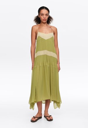 Kobieta ubrana w bezrękawową sukienkę midi w kolorze oliwkowo-zielonym i beżowym z cienkimi ramiączkami, plisowanymi detalami oraz czarnymi sandałami typu thong, stojąca na białym tle.