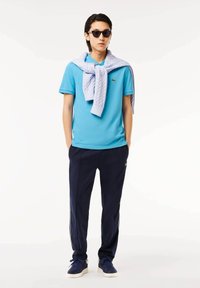Camisa de polo azul de manga corta, pantalones deportivos navy y zapatillas azules. Suéter de punto azul claro drapeado sobre los hombros, gafas de sol negras.