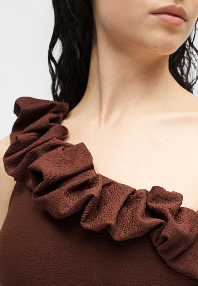 Κοντινό πλάνο γυναίκας που φοράει σκούρο καφέ τοπ με μια τιράντα και υφή, με μεγάλα υφασμάτινα βολάν κατά μήκος της γραμμής του λαιμού.