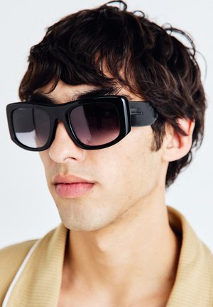 Gafas de sol negras rectangulares con lentes oscuros degradados, montura gruesa y acento de logo en el lateral, usadas por una persona con cabello despeinado.