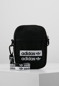 Černá látková crossbody taška s nastavitelným popruhem. Obsahuje bílé logo Adidas a značkové nášivky, má hladký povrch a zipové zavírání.