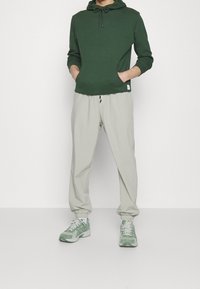 Person iført en mørkegrøn hættejakke, lysebeige bukser med elastiske manchetter og grønne og hvide sneakers, der står med hænderne i lommerne.