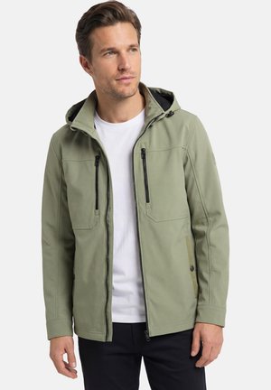 Mann trägt eine hellolivfarbene Kapuzenjacke über einem weißen T-Shirt und schwarzen Hosen und blickt leicht zur Seite.