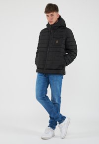 Giubbotto imbottito nero con zip frontale, cappuccio e motivo a cuciture orizzontali. Include una tasca sul petto con dettaglio del logo. Indossato con jeans blu e sneakers bianche.