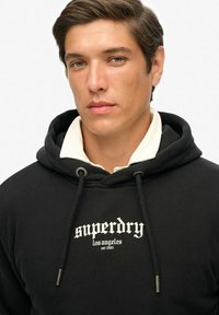 Felpa nera con cappuccio in tessuto morbido, con logo bianco "Superdry", coulisse con punte metalliche e colletto chiaro a contrasto.