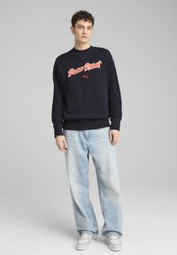 Sudadera navy con el texto "Puma Resort" en naranja, con puños y dobladillo acanalados. Combinada con jeans de pierna ancha de lavado claro y zapatillas grises.