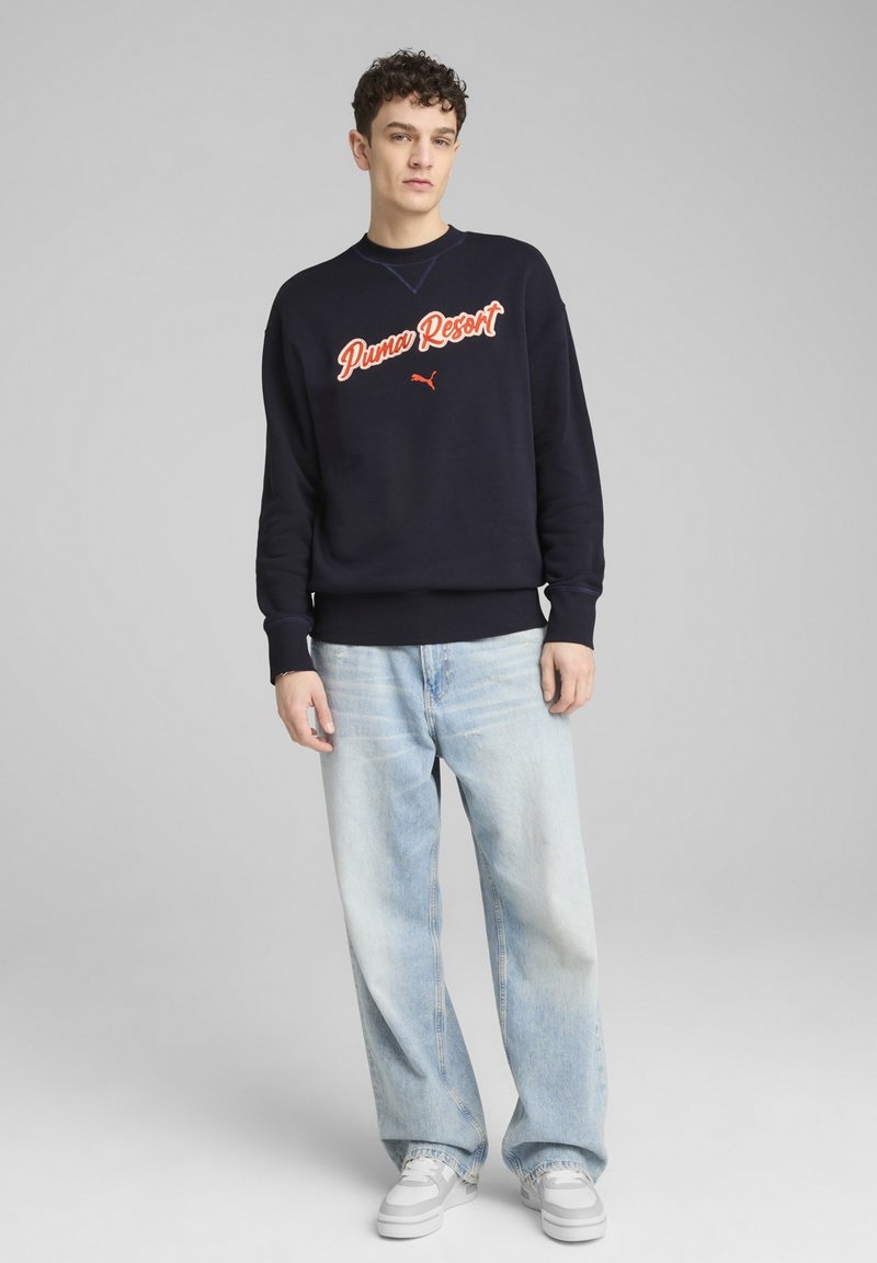 Sudadera navy con el texto "Puma Resort" en naranja, con puños y dobladillo acanalados. Combinada con jeans de pierna ancha de lavado claro y zapatillas grises.