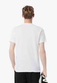 Joven con cabello corto castaño claro, vistiendo una camisa blanca de manga corta y pantalones negros, sosteniendo una toalla doblada en la mano derecha.