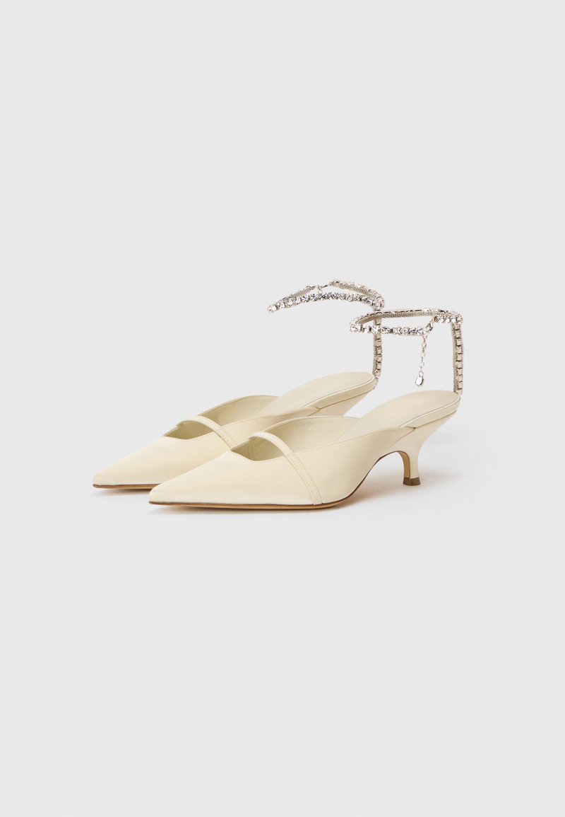 Cremefarvede spidse kitten heels med tynde stropper og rhinestone ankelarmbånd på hvid baggrund.