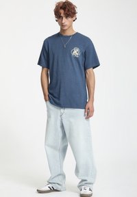 Camiseta de algodón azul con mangas cortas, cuello redondo y un estampado gráfico en el lado izquierdo del pecho. Pantalones vaqueros claros de corte holgado y zapatillas.