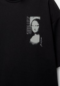 Schwarzes Baumwoll-T-Shirt mit einem bedruckten Bild der Mona Lisa und weißem Text, zentriert auf der Vorderseite. Lockerer Schnitt und kurze Ärmel.