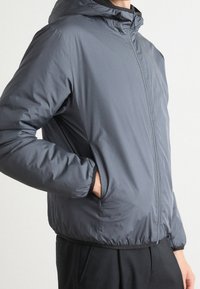 Grå huvjacka med en slät, lätt textur. Har en framzipper och elastiska muddärmar, med en avslappnad passform och sidofickor.