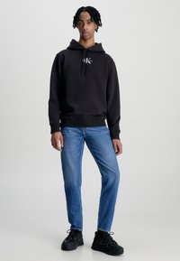 Felpa nera con cappuccio e cordino, con un piccolo logo sul petto. Indossata con jeans blu e sneakers nere. Vestibilità rilassata, design semplice.