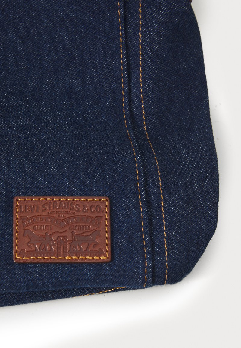 Blauwe denim stof met contrasterende oranje stiksels, voorzien van een rechthoekig bruinen leren label met reliëfbranding en decoratieve elementen.
