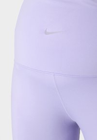 Světle fialové legíny mají hladkou texturu, středně vysoký pas a šedé logo Nike na levé straně. Šití je viditelné podél švů.