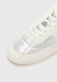 Baskets blanches avec un panneau latéral en métal argenté, bout rond et un design classique à lacets avec des détails texturés sur le matériau.