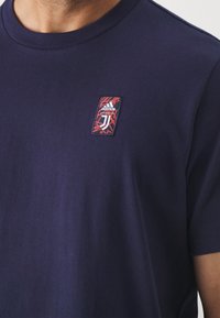 Námořnická modrá bavlněná tričko s kulatým výstřihem, s obdélníkovou nášivkou v červené, černé a bílé barvě, na které jsou loga Adidas a Juventus.