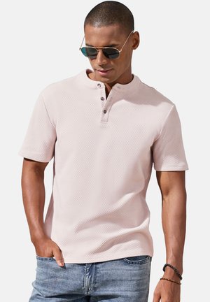 Mann trägt hellrosa kurzärmeliges, strukturiertes Henley-Shirt, blaue Jeans, Sonnenbrille und Armbänder, posiert mit einer Hand in der Hosentasche.