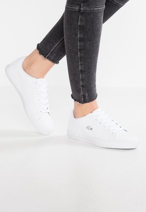 Personne portant des baskets blanches à lacets avec un logo sur le côté et un jean noir à ourlet effiloché sur fond blanc.