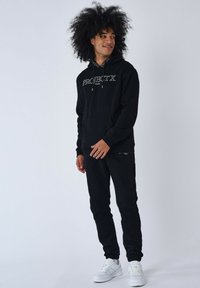 Sudadera negra con el texto "PROJECTX", con un bolsillo frontal y capucha ajustable, combinada con joggers negros a juego y zapatillas blancas.